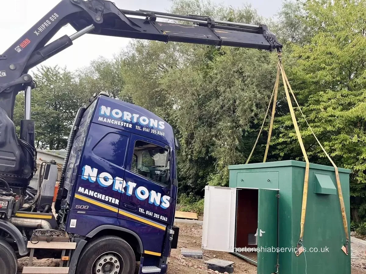 A Nortons rigid with its crane lifting a green kiosk unit beside a lorry with an empty bed