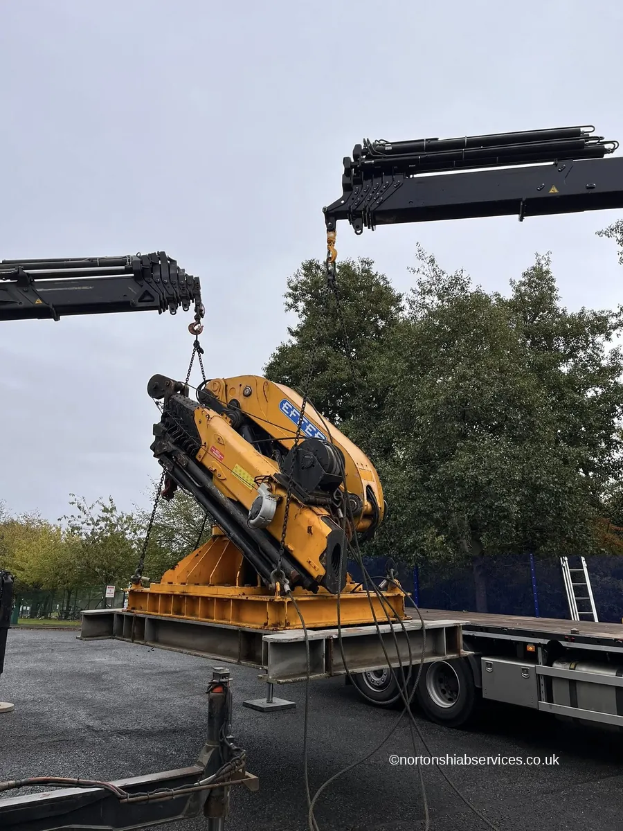 A yellow lorry loader crane being lifted onto a Nortons flatbed beside a site cabin