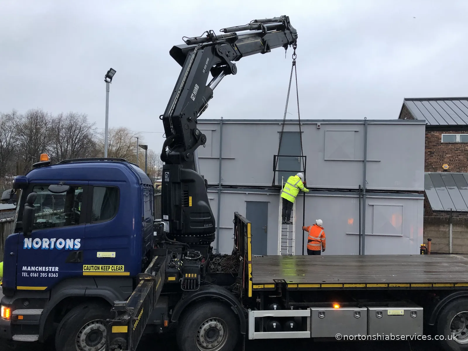 Two operatives on a ladder checking a run of grey portable cabins at the Nortons yard, with the lorry crane slung above