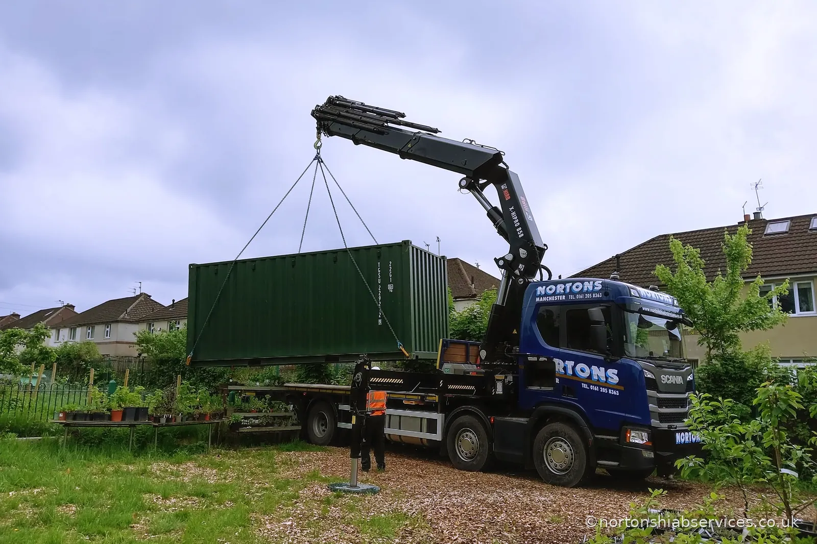 A refurbished portable cabin slung on the crane over a garden fence from a Nortons eight wheeler