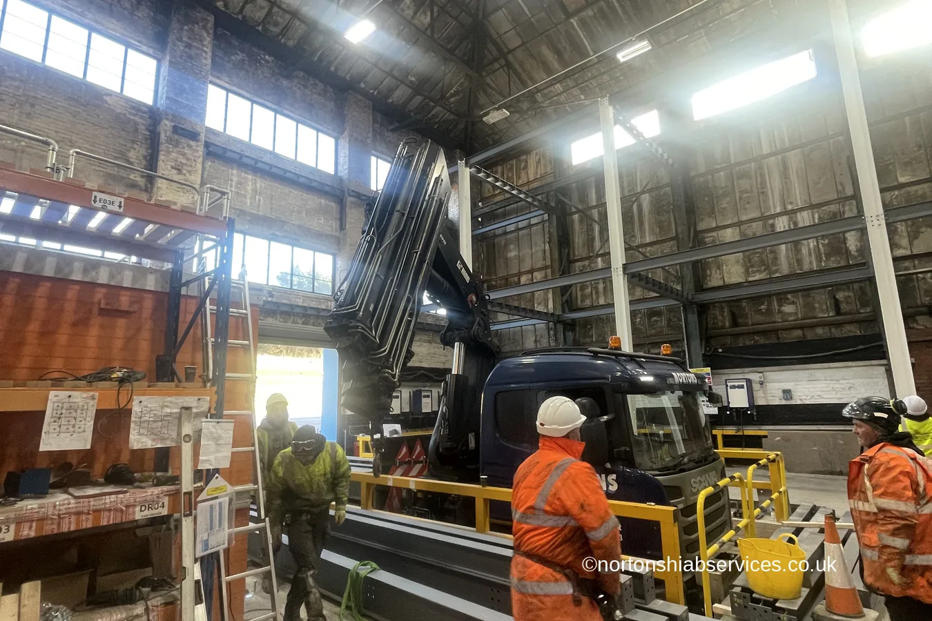 A Nortons lorry crane inside a factory building lifting steel beams, with operatives in high-visibility clothing alongside