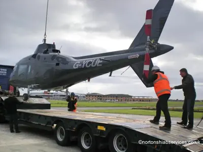 Two men steadying the suspended helicopter as it is guided over the trailer deck