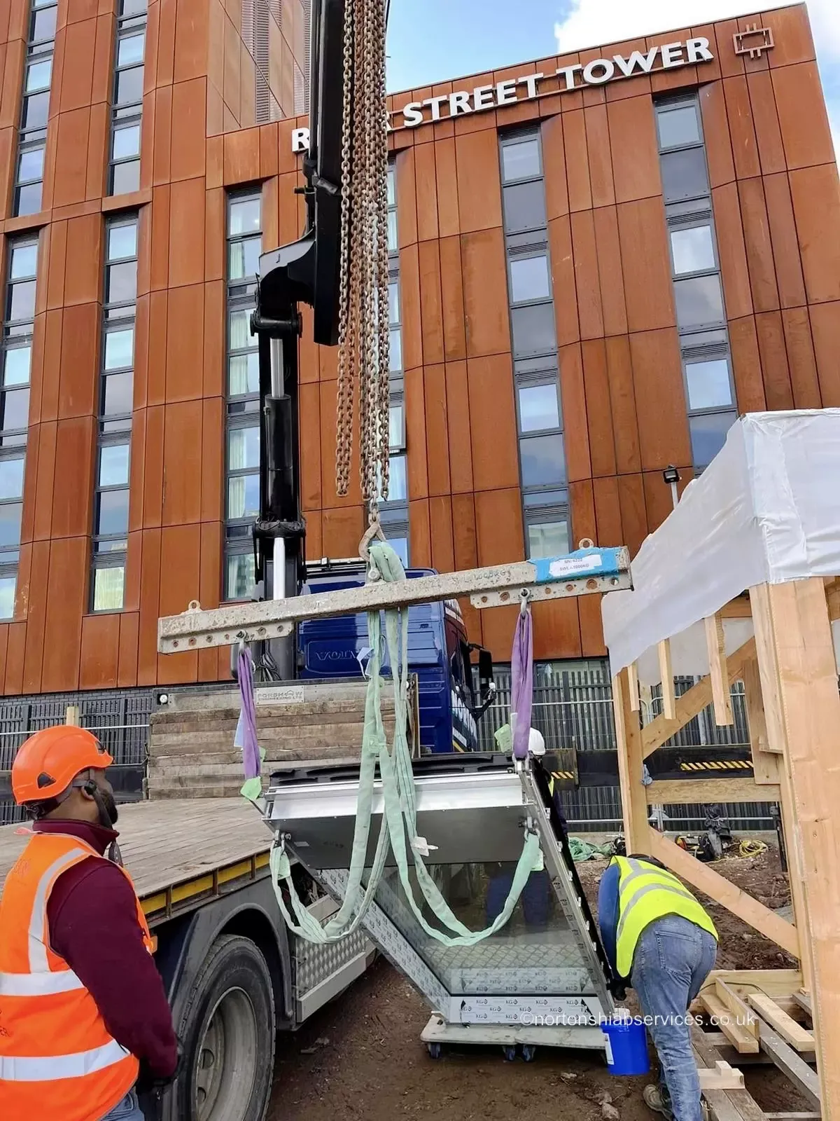 A Nortons crane lifting a steel frame between a tower block and a hoarding on a city street