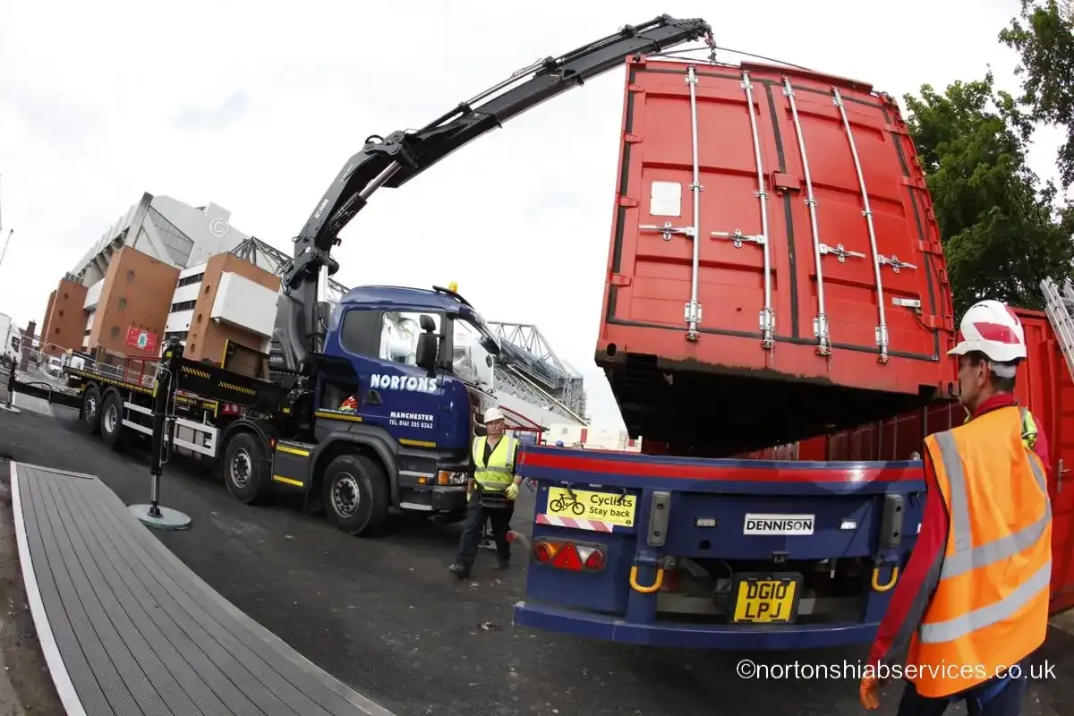 A Nortons crane lifting a red site container clear of a trailer beside a stadium hoarding