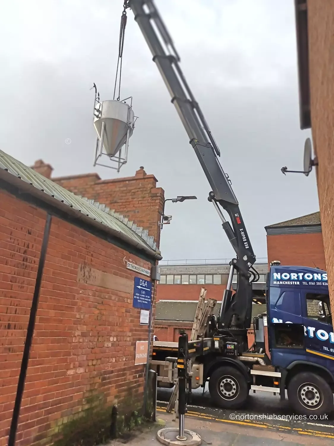A Nortons hiab lifting a stainless steel brewing vessel in from the street, clear of a brick yard wall