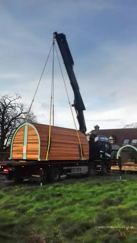 A Nortons lorry crane lowering a timber glamping pod onto a grass field