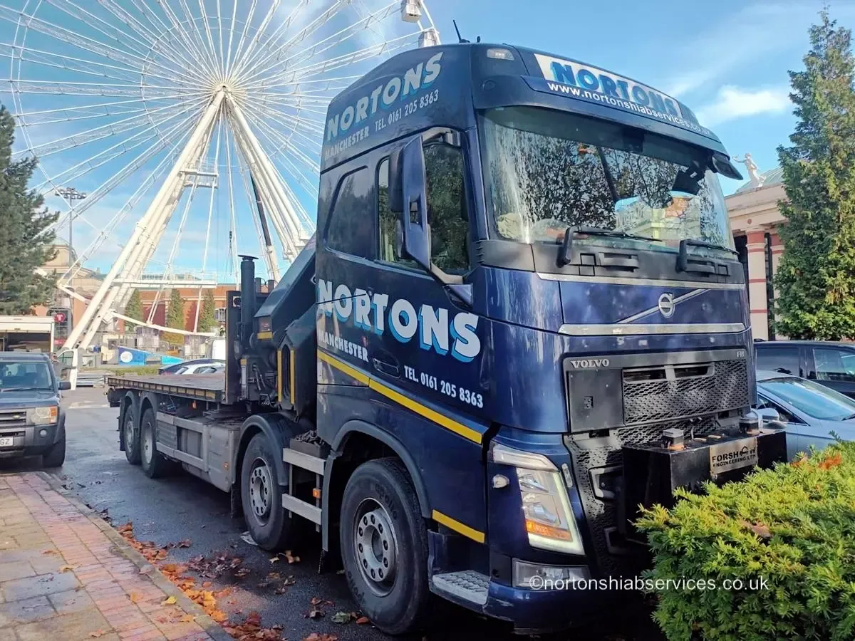 A Nortons Volvo rigid with an empty flat bed parked on a city street beside a large wheel
