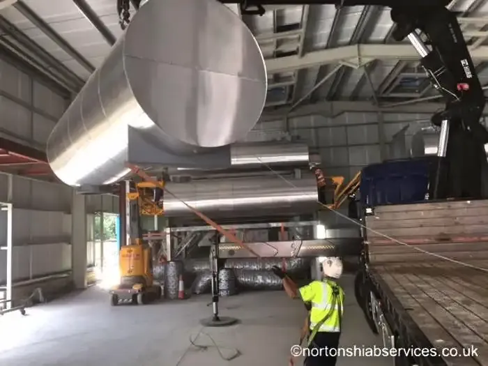Large stainless ductwork lifted overhead inside a building with a banksman below