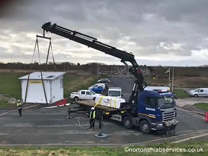 A Nortons crane lifting a small lifeguard kiosk from the lorry bed onto open ground beside a road