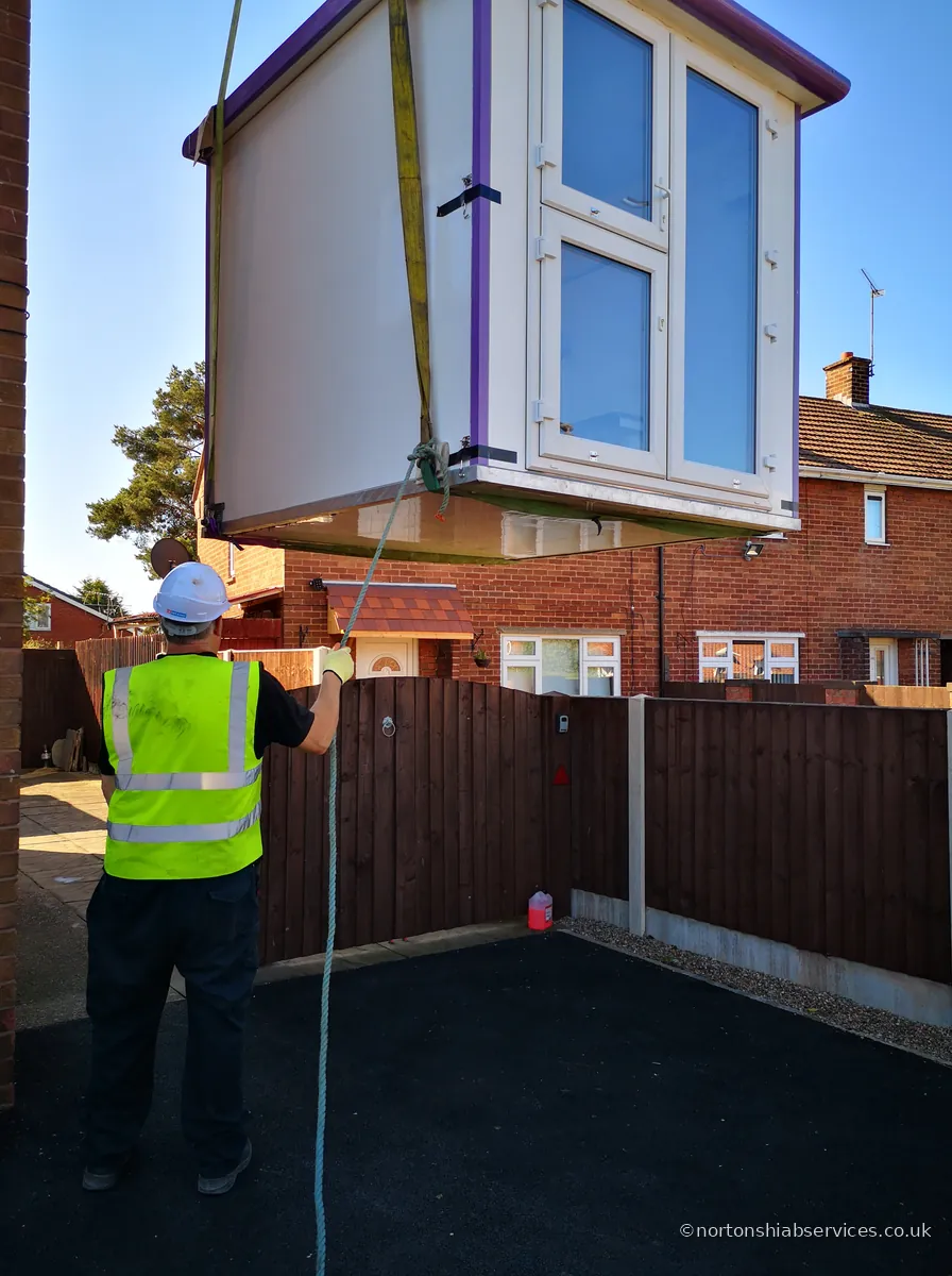 A Nortons crane lifting a glazed garden room over a fence into a back garden