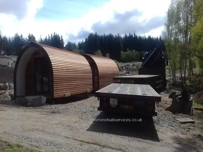 Two cedar clad glamping pods on prepared bases at a wooded site beside an empty Nortons trailer