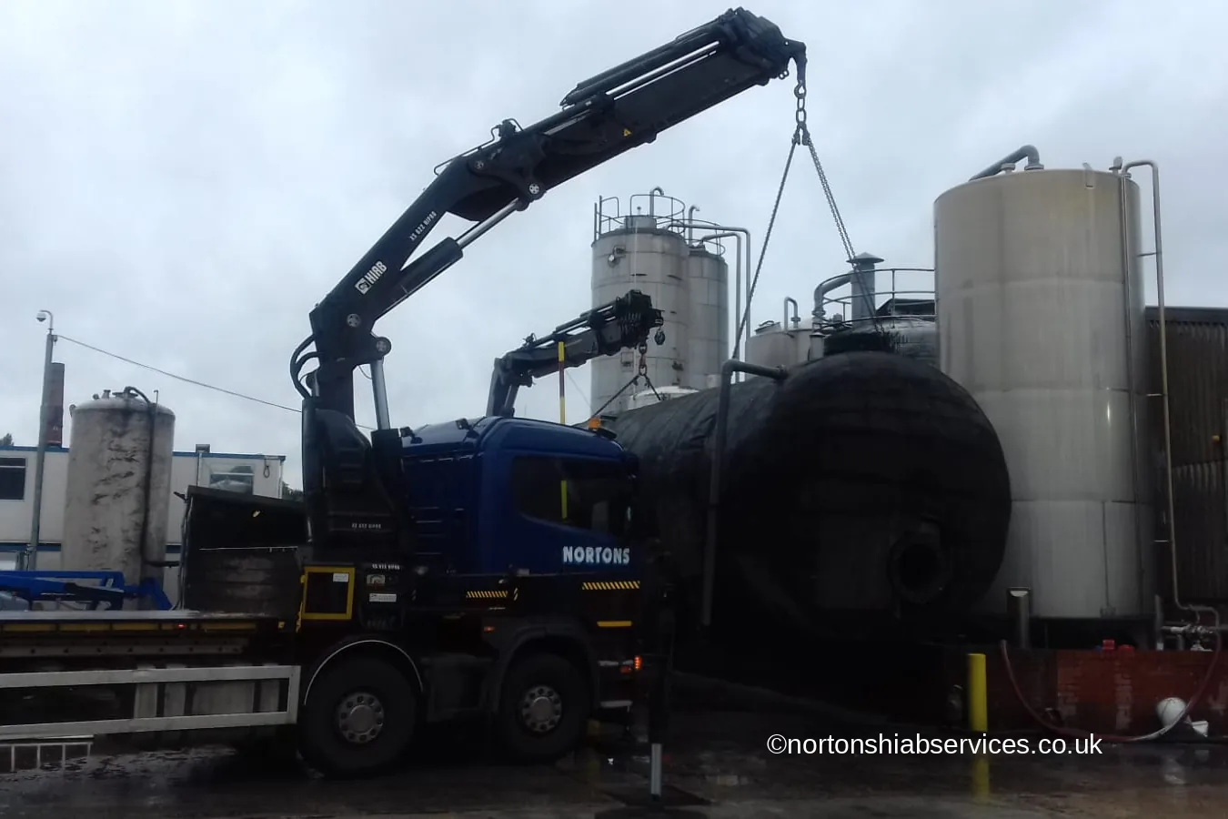 A Nortons hiab lifting a black tank inside a working tank farm