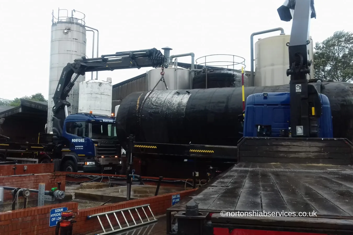 A Nortons hiab lifting large black process tanks inside a working chemical plant