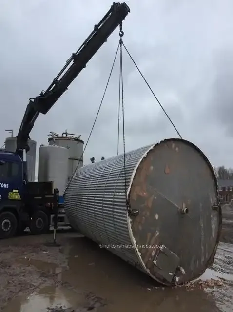 A Nortons crane lowering a tall corrugated steel tank onto its side in a compound