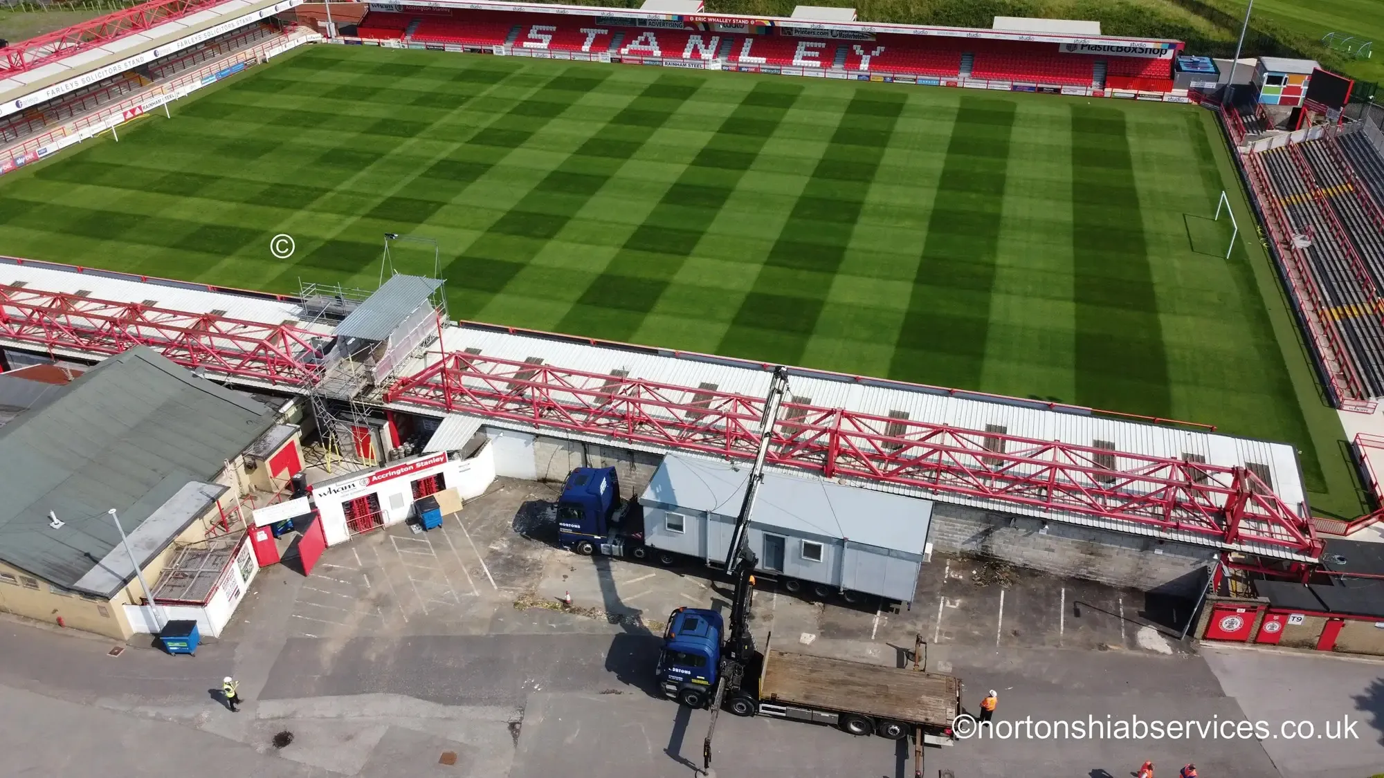 An aerial view of Accrington Stanley's ground with red roof trusses laid out beside the stand and Nortons hiab vehicles working