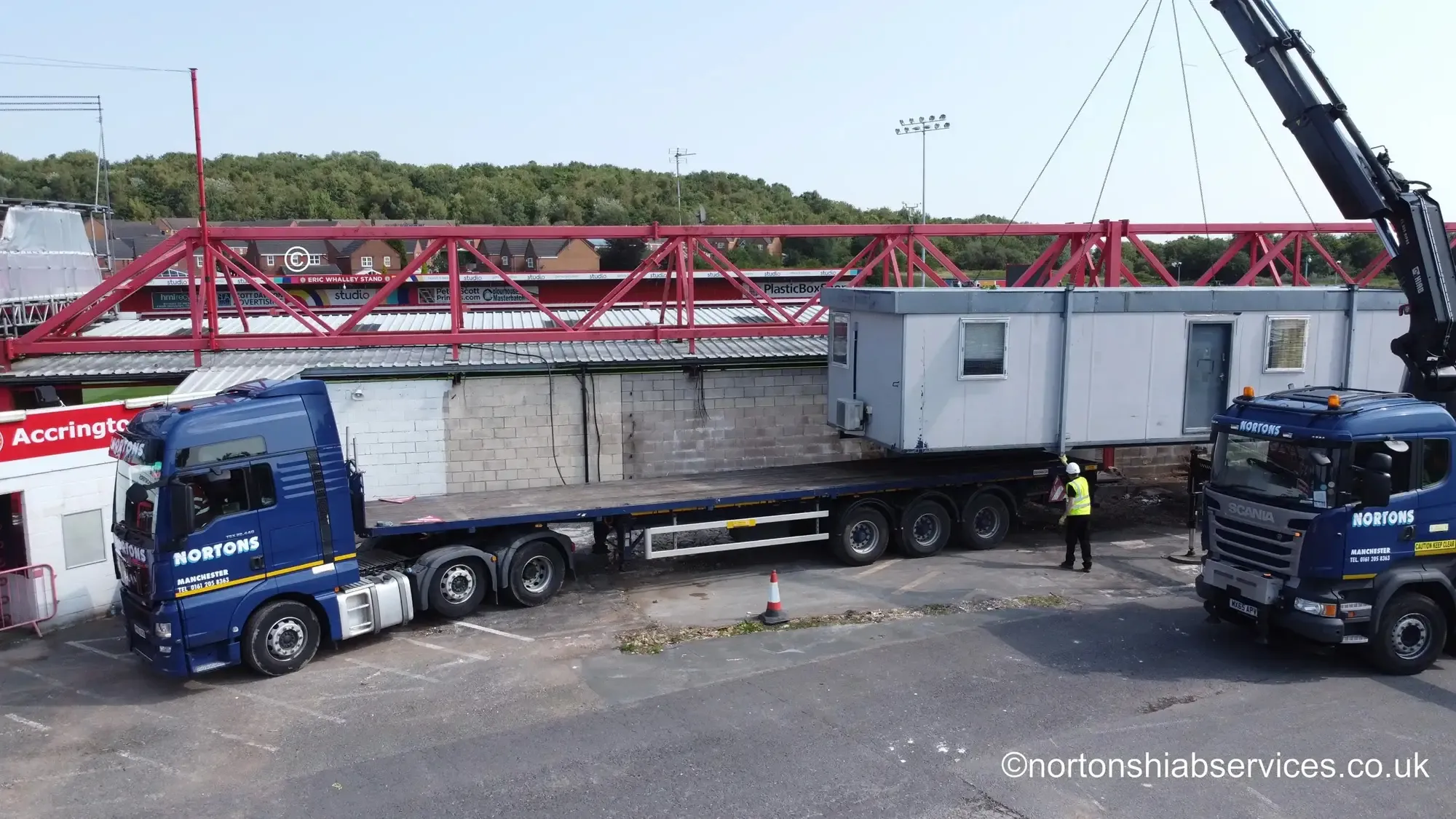 Red steel roof trusses laid out beside a Nortons artic and a rigid craning a site cabin at a football ground