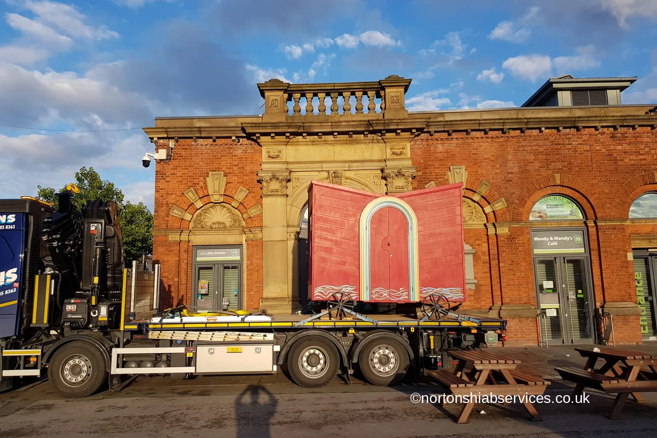 A red travelling arts unit set down by a Nortons crane outside a red brick museum