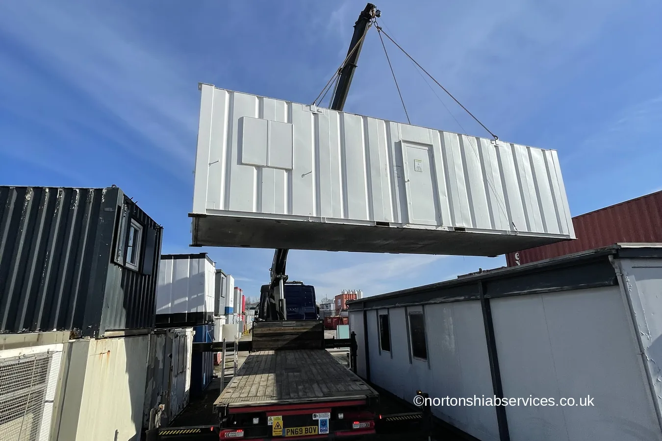 A portable cabin suspended above a yard by a Nortons hiab