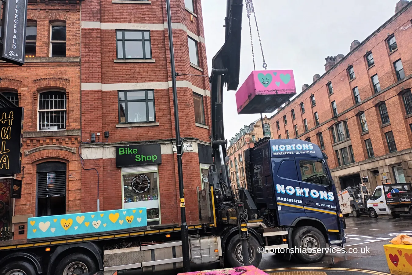 A Nortons crane lifting a painted concrete bollard on chains in a Manchester street