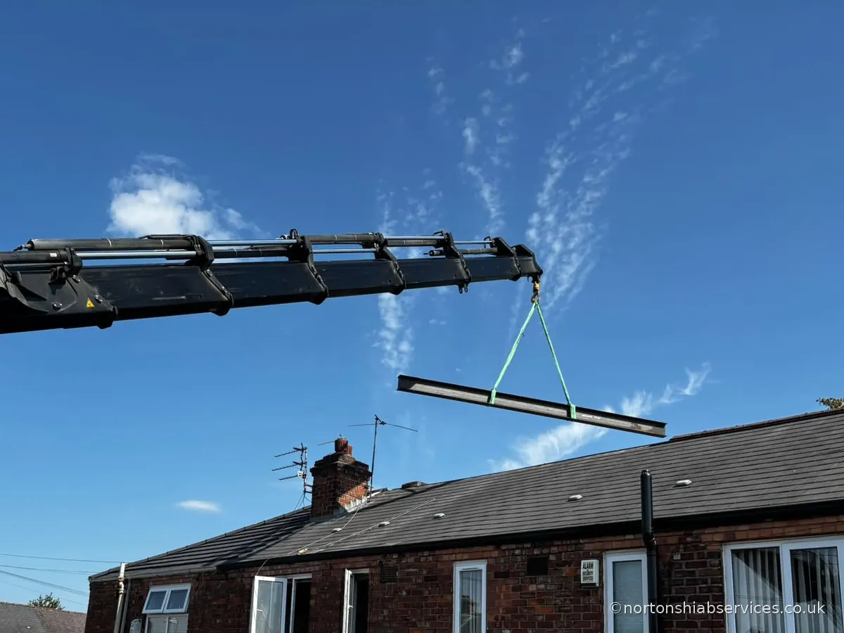 A Nortons hiab with the boom fully extended carrying a steel beam on green slings above a terraced roofline at Eccles