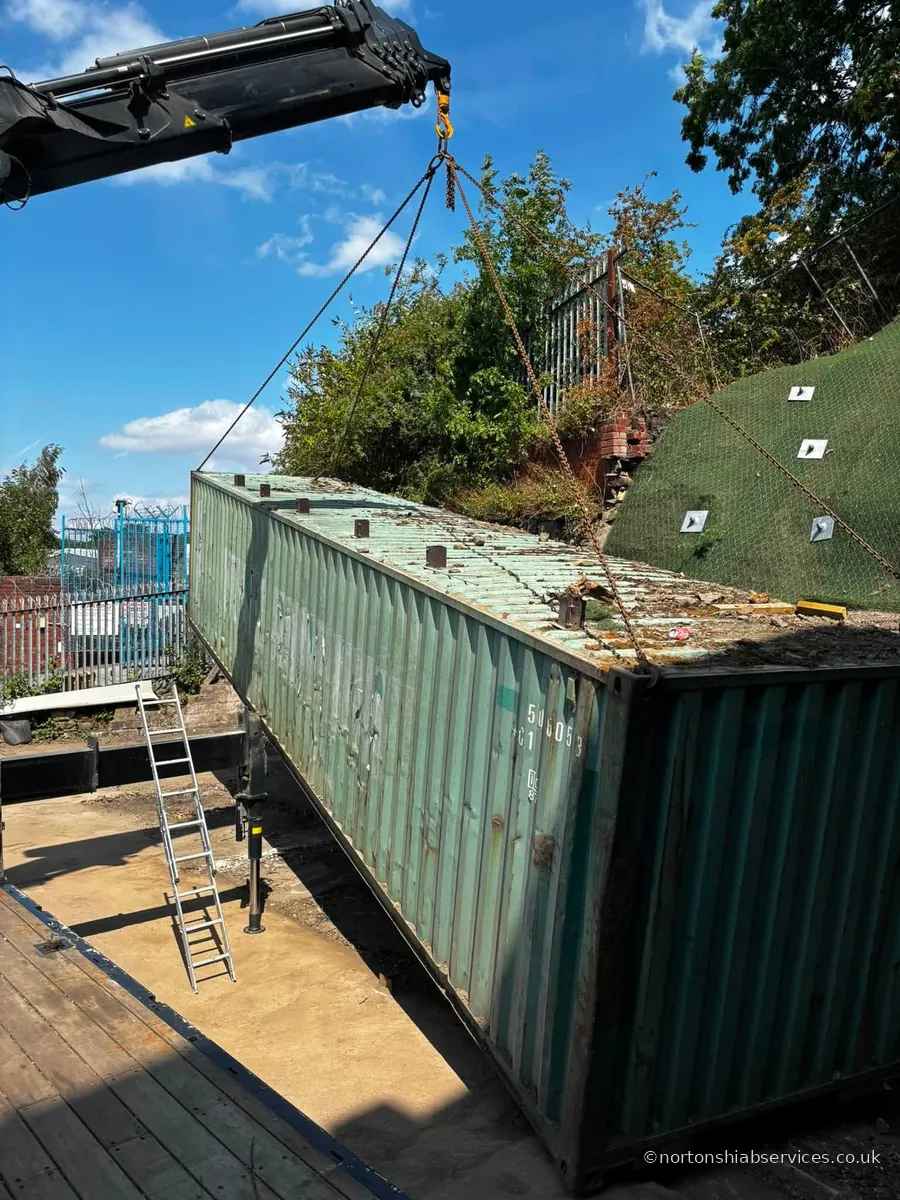 A Nortons crane lifting a green forty foot container off the lorry bed beside a netted embankment at Sheffield