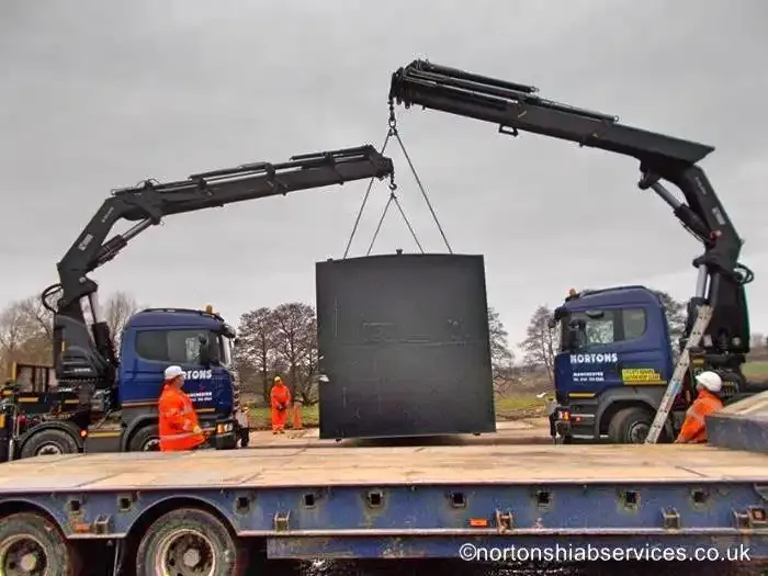 A Nortons lorry crane lowering a large black tank onto a low loader