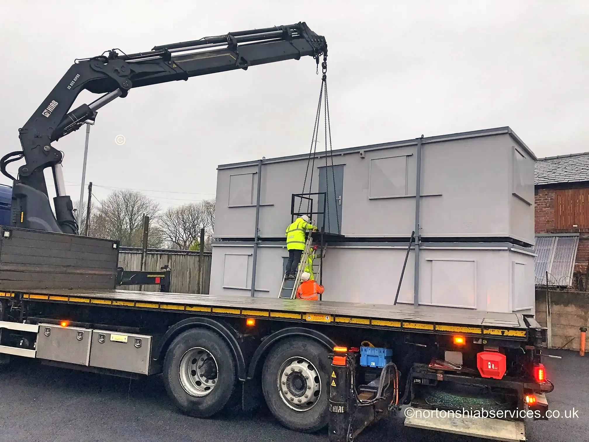 A Nortons lorry crane lifting a portable cabin with an operative on a ladder alongside