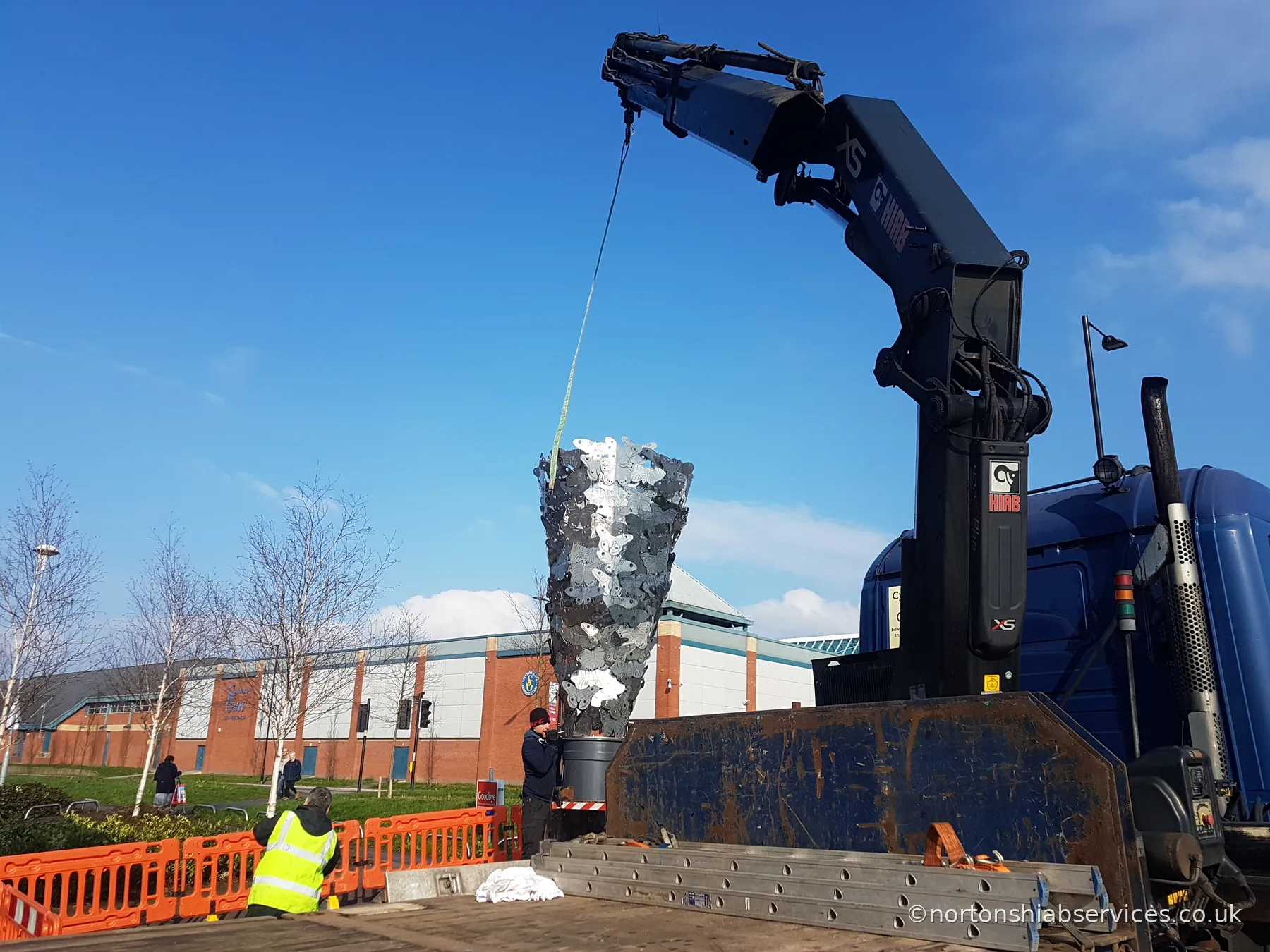 A Nortons lorry crane lowering a tall openwork metal sculpture onto its plinth against a clear blue sky