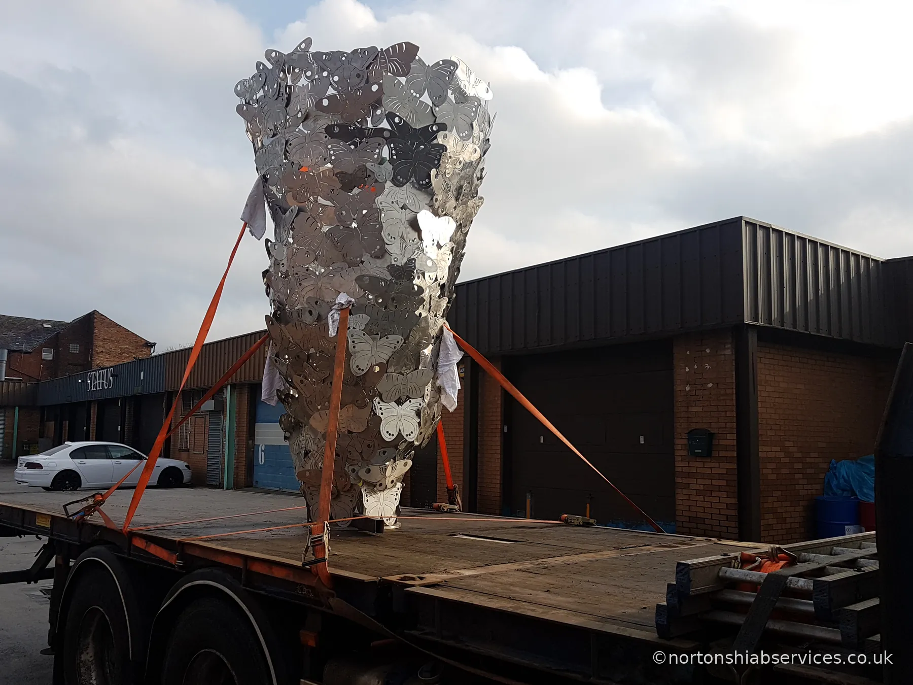 A tall openwork metal sculpture standing upright on a Nortons flatbed, held by four orange ratchet straps
