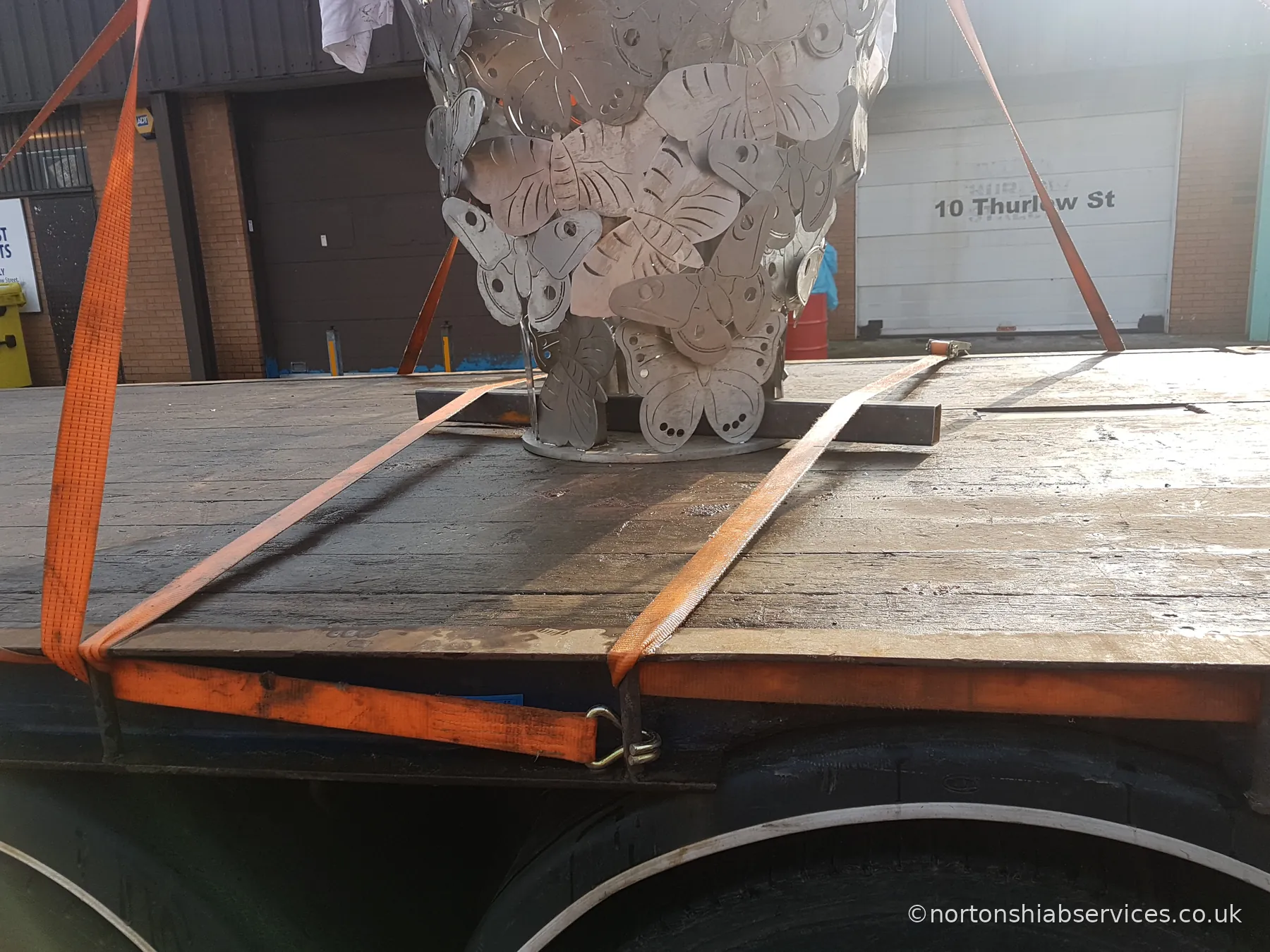 Close view of the base of a metal sculpture chocked with steel sections and held by orange ratchet straps on a lorry deck