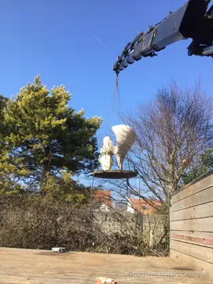 The white cast sculpture suspended on the crane away from the lorry with trees behind it