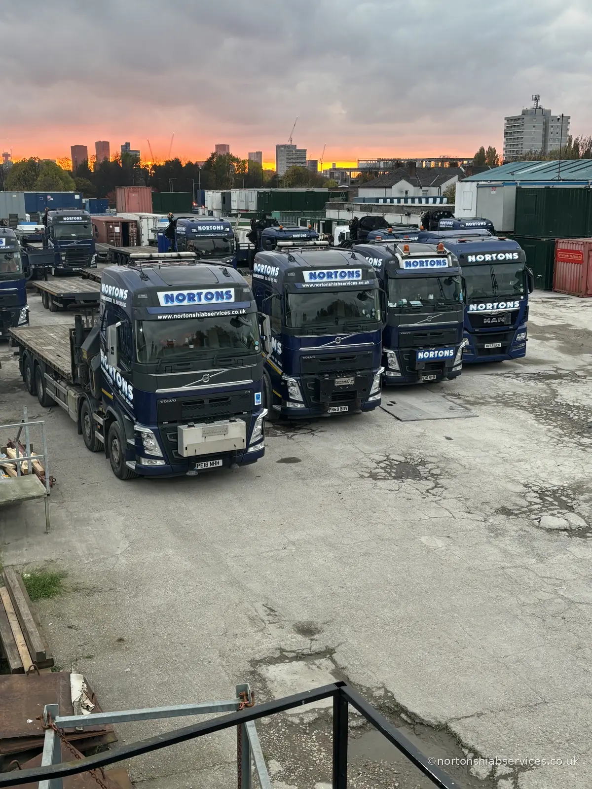 Nortons hiab lorries parked in the Manchester storage yard at dusk with containers and the city skyline behind