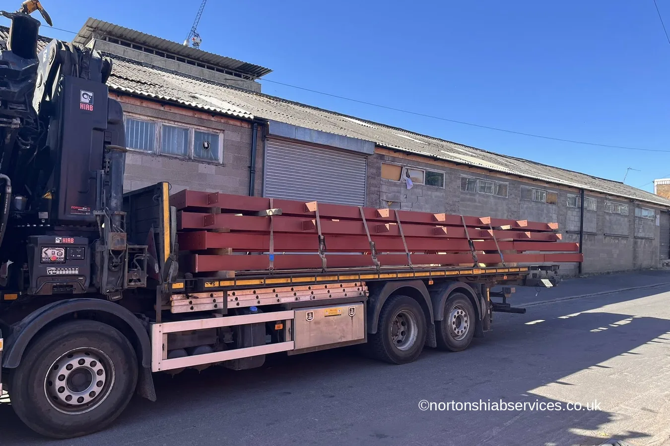 A Nortons rigid loaded with red painted steel sections standing alongside a long stone built industrial building