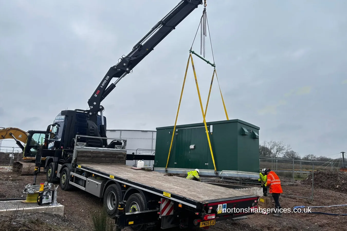 A Nortons crane lorry holding a dark green kiosk unit on yellow slings beside its prepared base at Winsford