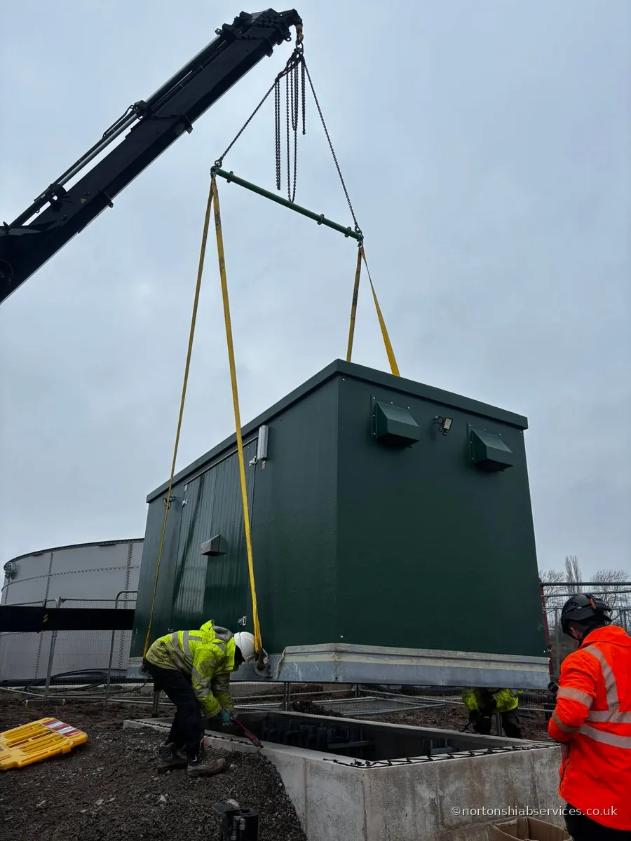 A Nortons crane lowering a dark green kiosk unit onto its concrete base at Winsford in Cheshire, with two banksmen guiding it in