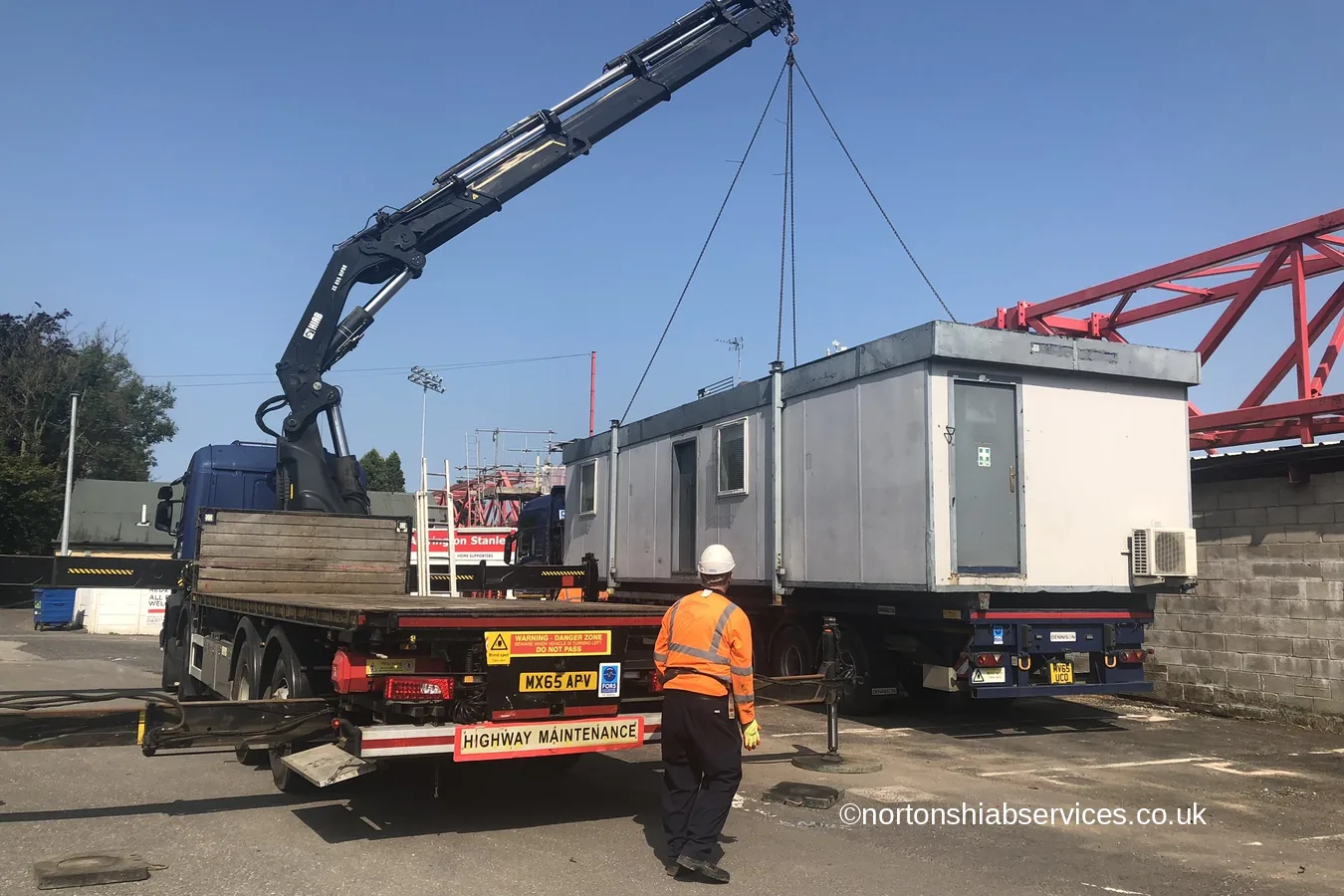 A Nortons crane lorry lifting a site cabin off the deck at Accrington while a banksman stands clear