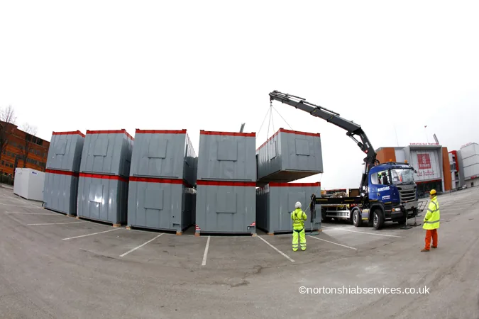A Nortons crane lorry taking a grey modular unit off a stack in a car park at the cricket ground in Trafford