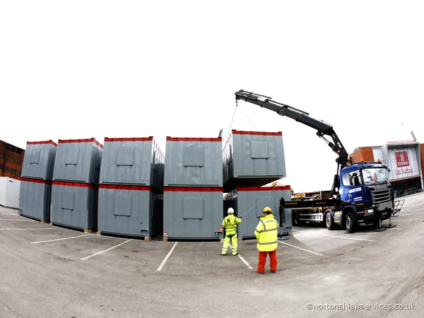 A Nortons hiab lifting large grey modular units into position in a car park at the cricket ground in Trafford, with two banksmen guiding them