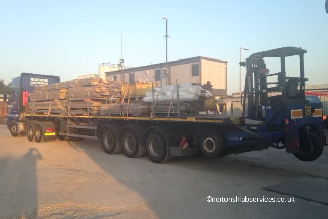 A Nortons trailer loaded with scaffolding beside a forklift and site cabins in a distribution yard