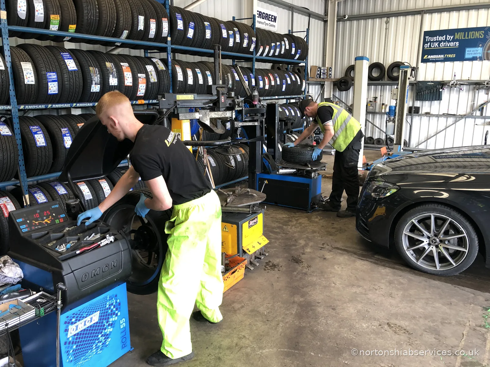 Two Nortons Tyres fitters working on wheels in the fitting bay, with racked tyres behind them and a car on the ramp