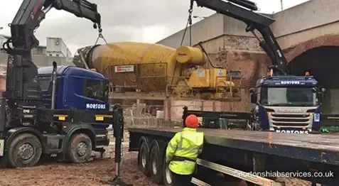 Two Nortons hiabs lifting a large yellow cement mixer drum with a banksman watching