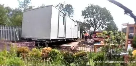 White portable cabin being set down from a Nortons vehicle beside a hedge