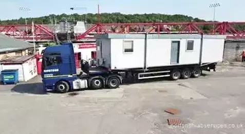 Long white cabin on a Nortons trailer in front of a football stand