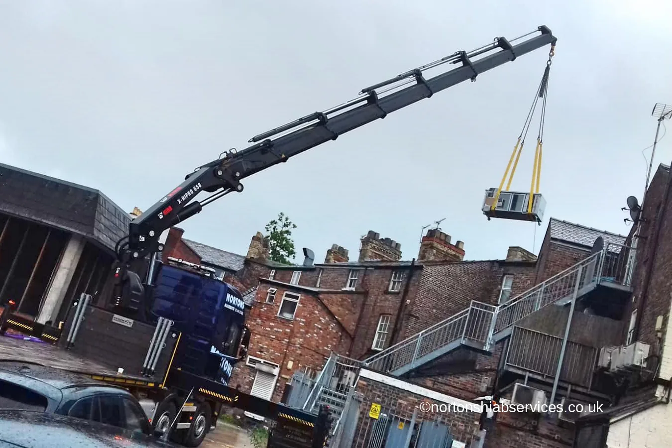 A Nortons lorry crane with its boom extended over the rooftops of a terrace, lowering an air conditioning unit into the yard behind