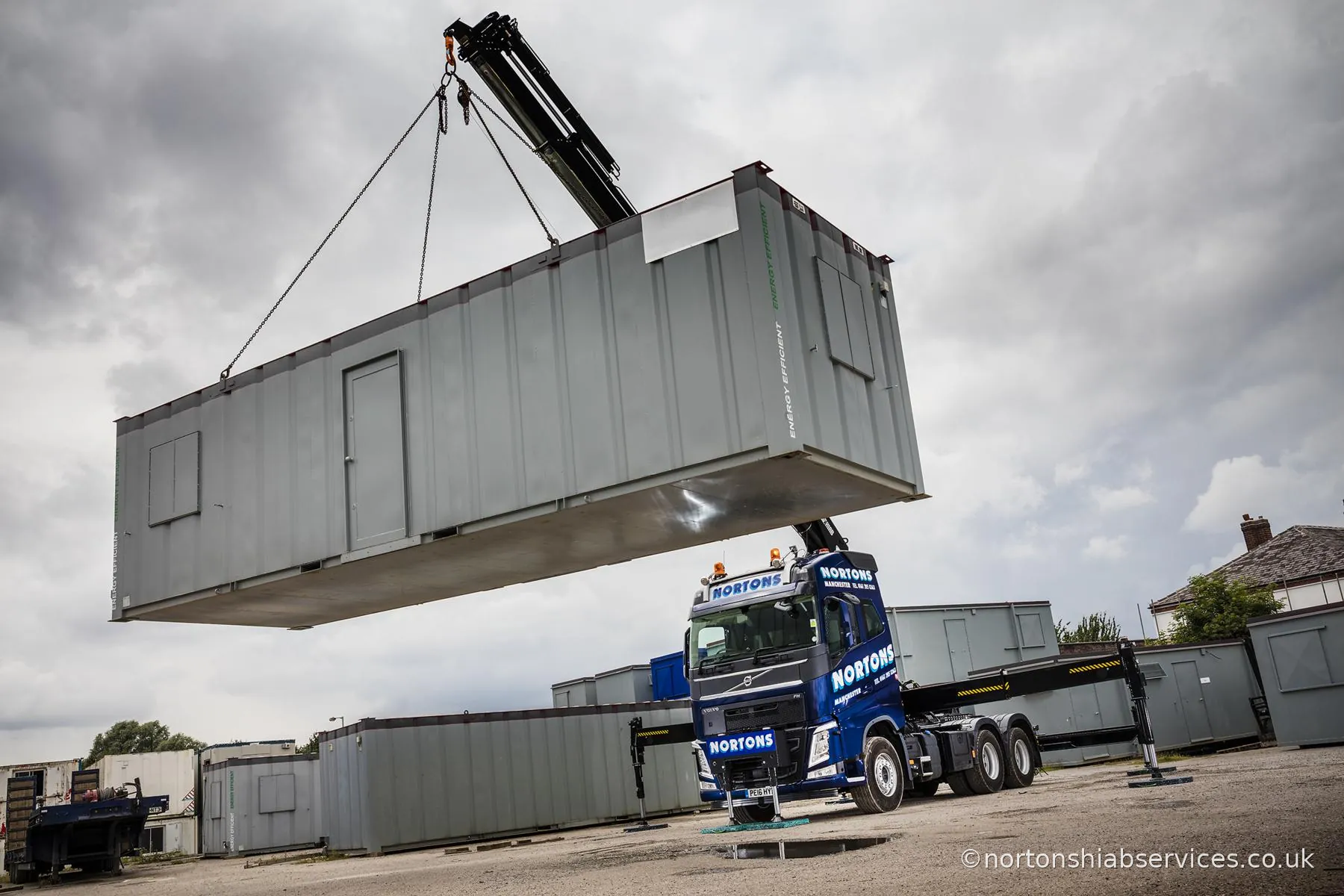 A Nortons lorry standing on its outriggers with a site cabin lifted high on chains